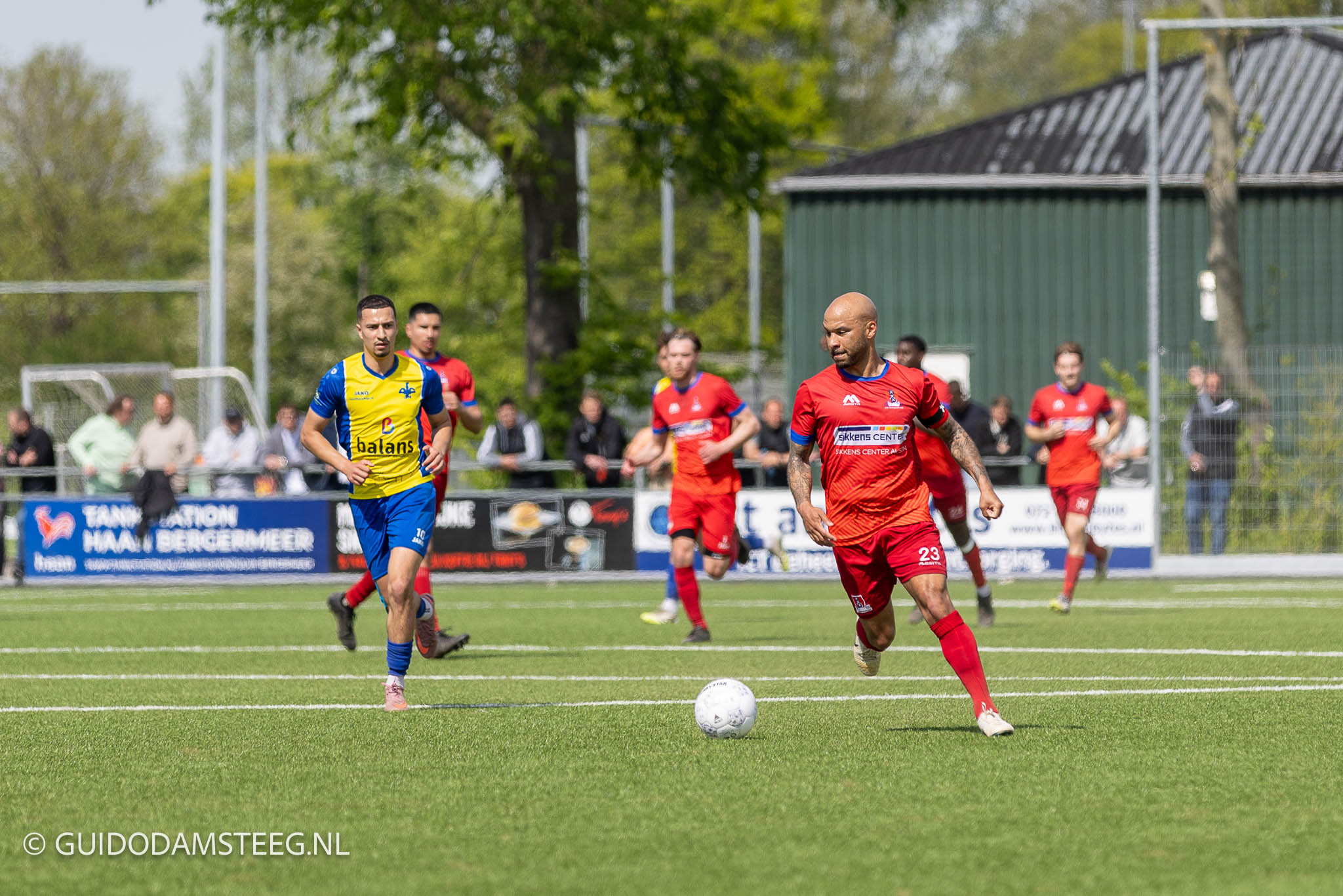 Soccer match on a sunny day with players in red and blue-yellow kits chasing the ball on a green field, spectators behind a fence in the background.