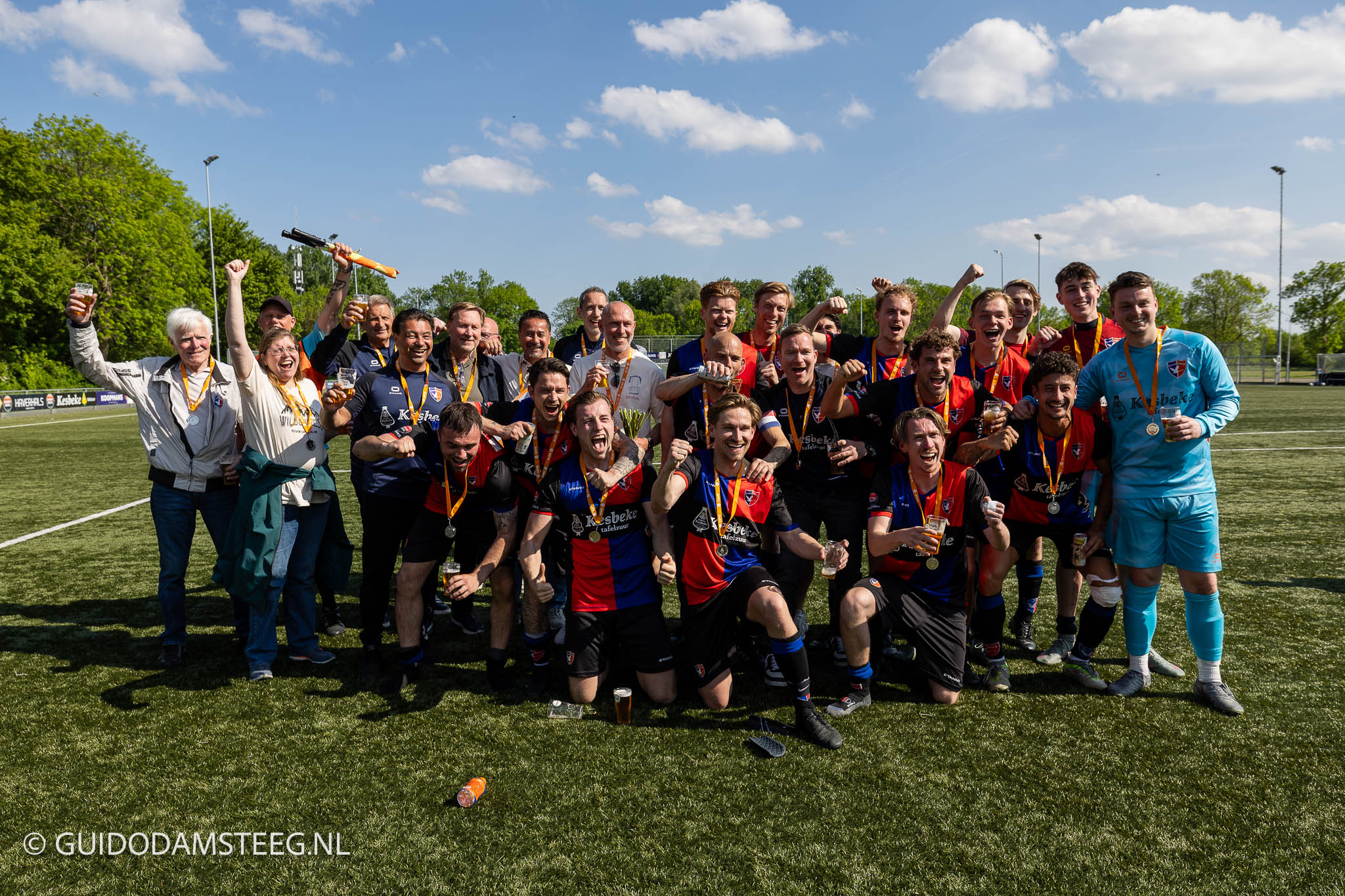 Large group of soccer players and supporters celebrate on a sunny field, wearing medals and holding drinks.