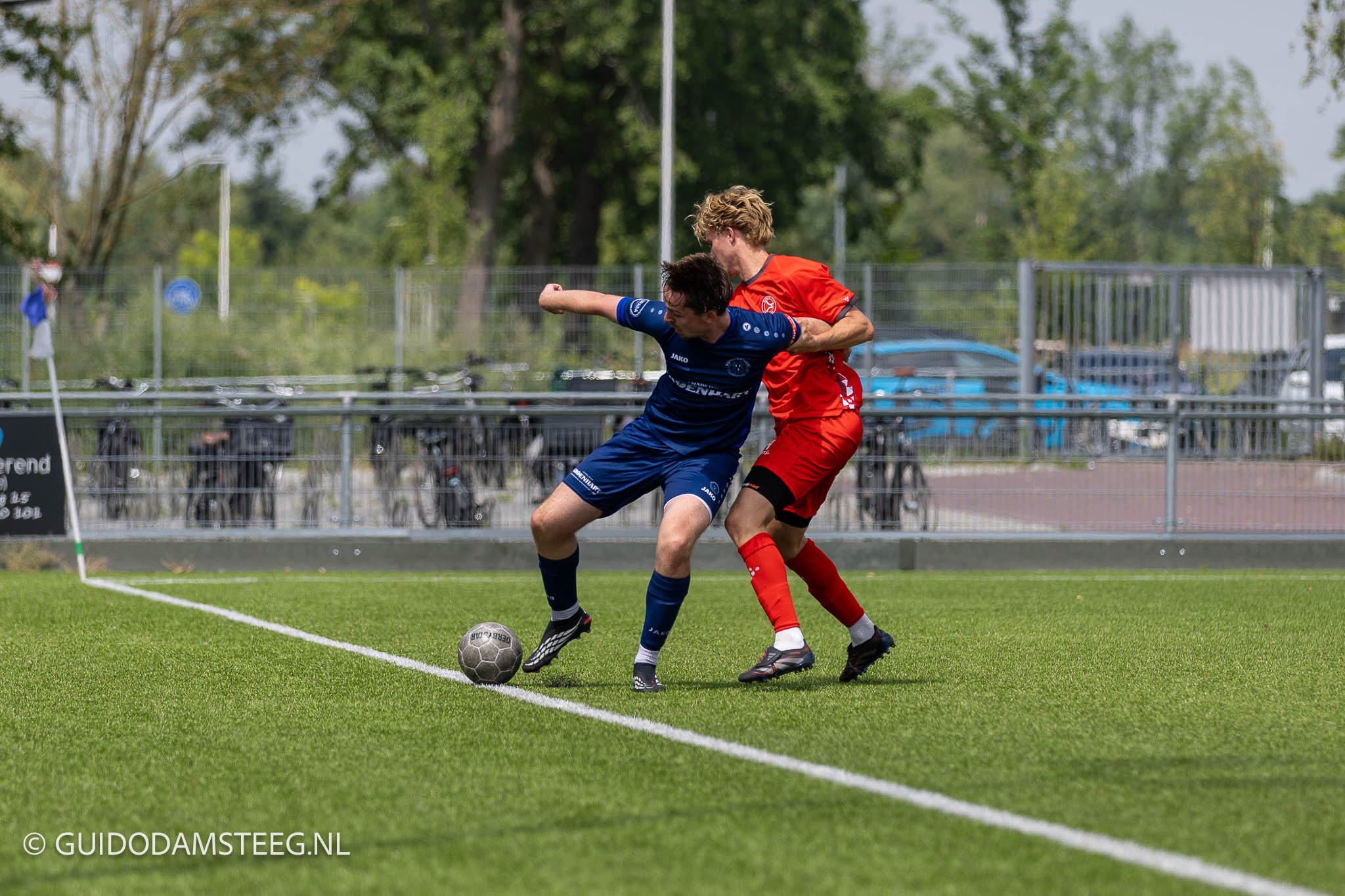Purmersteijn - Jong Almere City 2-1 11 juli 2026 - Guido Damsteeg - Guido Damsteeg Two soccer players in blue and red battle for the ball on a sunny grassy field, near a sideline with a fence and trees in the background in action. (Informative moment of contest for possession)